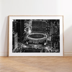 May include: Black and white framed print of a city at night. The image features a large, circular building surrounded by skyscrapers and city lights. The print is displayed on a wooden floor.