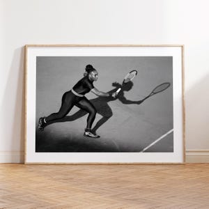 May include: Black and white framed print of a tennis player in action, swinging a racket on a court. The player is wearing a black outfit and athletic shoes. The image captures the player's shadow and the court's lines.