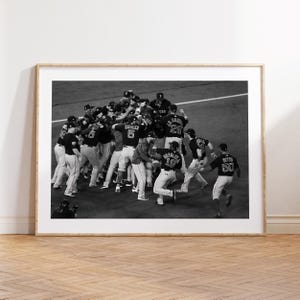 May include: Black and white framed photograph of a baseball team celebrating a victory. Players in uniforms are huddled together, with some running towards the group. The image is set against a backdrop of a baseball field.