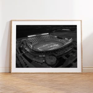 以下が含まれることがあります： 夜にライトアップされたスタジアムの白黒航空写真。画像には、観客席と競技場が表示されています。写真は明るい木製のフレームに入っています。