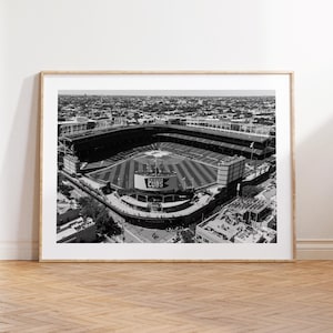 Puede incluir: Impresión enmarcada en blanco y negro del Wrigley Field, hogar de los Chicago Cubs. La vista aérea muestra el estadio de béisbol con el marcador icónico y la ciudad circundante. La impresión se muestra sobre un suelo de madera.