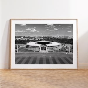Könnte beinhalten: Schwarz-Weiß-Druck in einem Rahmen eines großen Stadions mit kreisförmigem Design. Das Stadion hat ein großes offenes Feld davor mit gestreiftem Gras. Der Himmel ist voller Wolken.