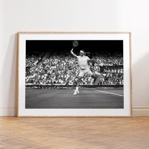 May include: Black and white framed print of a tennis player mid-swing, leaping in the air with a racket. The player is wearing a white outfit and headband. A crowd of spectators fills the background.