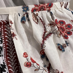 May include: Close-up of a white fabric with a floral pattern in red, blue, and brown. The fabric appears to be part of a skirt or dress, with a gathered waistband visible at the top. The design features stylized flowers and leaves.