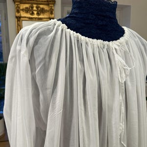 May include: A white, flowing garment with a gathered neckline and long sleeves. The neckline features a dark blue lace collar. The fabric appears lightweight and semi-sheer, with a drawstring detail at the neck.