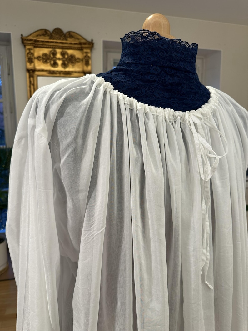 May include: A white, flowing garment with a gathered neckline and long sleeves. The neckline features a dark blue lace collar. The fabric appears lightweight and semi-sheer, with a drawstring detail at the neck.