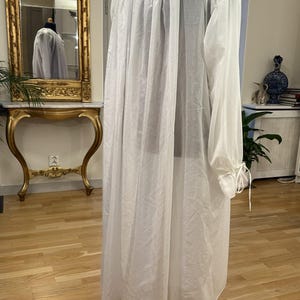 May include: A long, white, sheer garment with long sleeves and a high, gathered neckline. The sleeves are gathered at the wrists with ties. The garment is displayed on a wooden mannequin. A mirror and antique furniture are in the background.