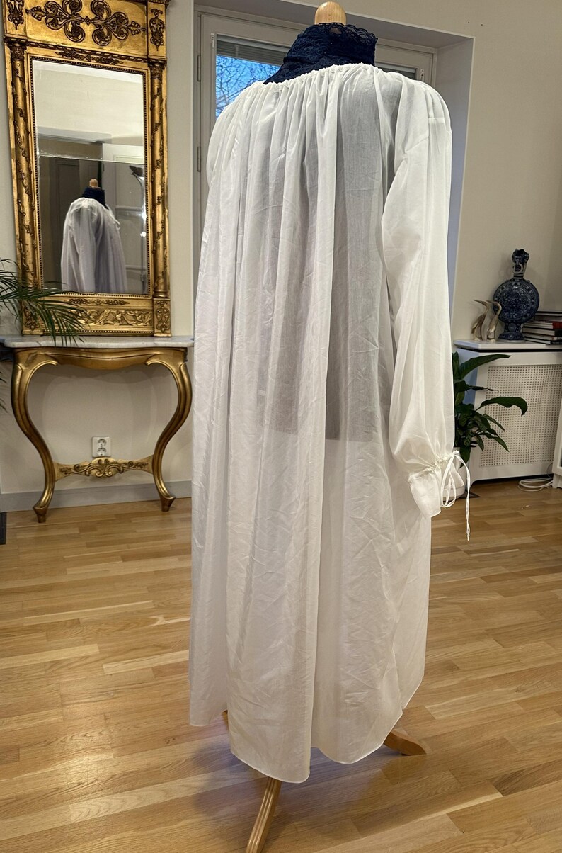 May include: A long, white, sheer garment with long sleeves and a high, gathered neckline. The sleeves are gathered at the wrists with ties. The garment is displayed on a wooden mannequin. A mirror and antique furniture are in the background.