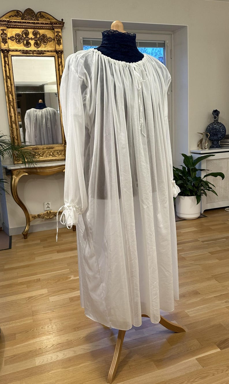 May include: A long, white, sheer nightdress with long sleeves and a high, dark collar. The sleeves are gathered at the wrists with ties. The nightdress is displayed on a wooden dress form. The garment is likely vintage or inspired by historical styles.