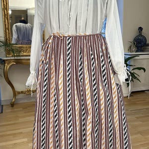 May include: A vintage outfit on a mannequin. It features a white blouse with long sleeves and a drawstring neckline, paired with a long skirt. The skirt has vertical stripes in brown, black, and gold. The outfit is displayed on a wooden stand.