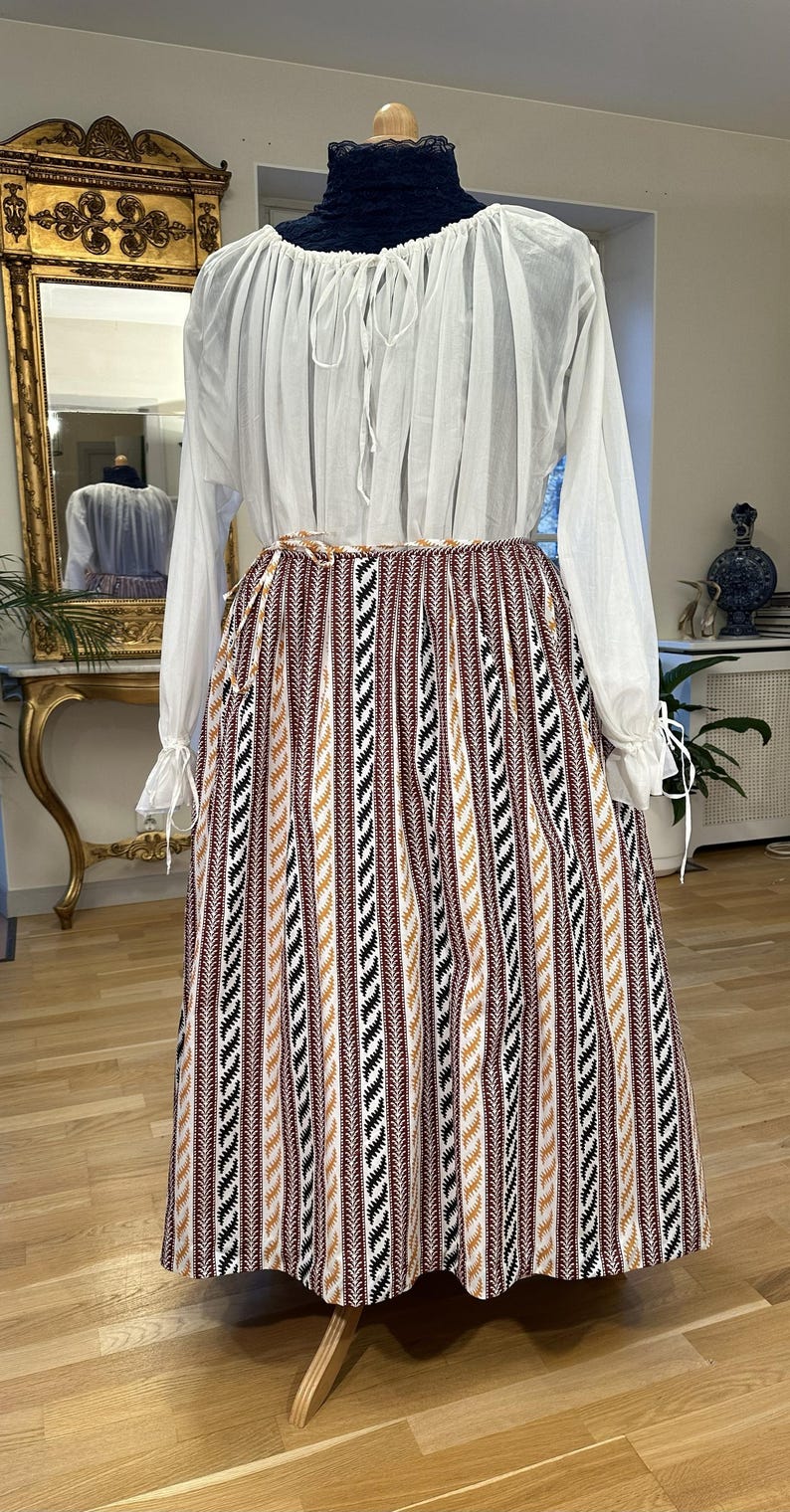 May include: A vintage outfit on a mannequin. It features a white blouse with long sleeves and a drawstring neckline, paired with a long skirt. The skirt has vertical stripes in brown, black, and gold. The outfit is displayed on a wooden stand.