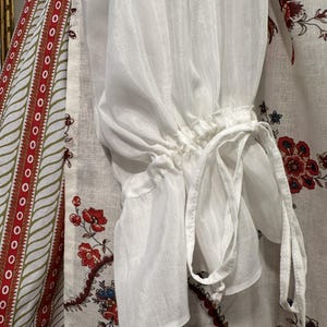 May include: Close-up of a white, sheer blouse sleeve with a gathered cuff and tie detail. The sleeve is made of a lightweight fabric. The background features a patterned fabric with red and blue floral designs.