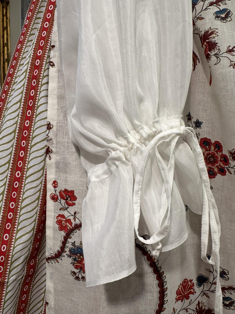 May include: Close-up of a white, sheer blouse sleeve with a gathered cuff and tie detail. The sleeve is made of a lightweight fabric. The background features a patterned fabric with red and blue floral designs.