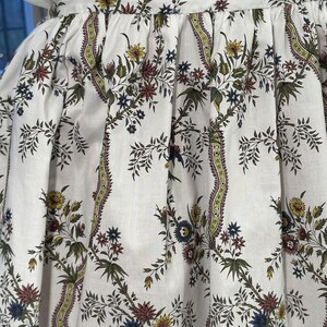 May include: White fabric with a floral pattern in yellow, blue, red, and green. The design features stylized flowers and foliage, creating a vintage aesthetic. The fabric appears to be part of a skirt or dress.