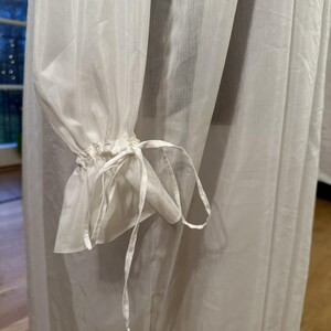 May include: Close-up of a white, sheer fabric garment. The sleeves are gathered at the wrist with a delicate tie detail. The fabric appears lightweight and flowing, suggesting a dress or blouse.