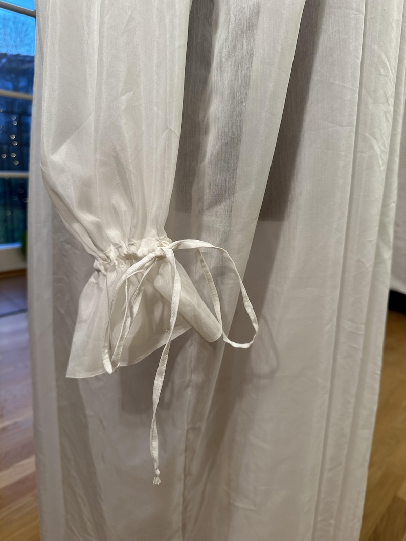 May include: Close-up of a white, sheer fabric garment. The sleeves are gathered at the wrist with a delicate tie detail. The fabric appears lightweight and flowing, suggesting a dress or blouse.