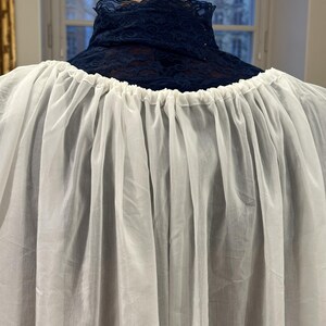 May include: A white, gathered fabric garment with a high, navy blue lace collar. The garment is displayed on a wooden mannequin head, showcasing the texture and drape of the fabric. The background includes a window with a view of buildings.