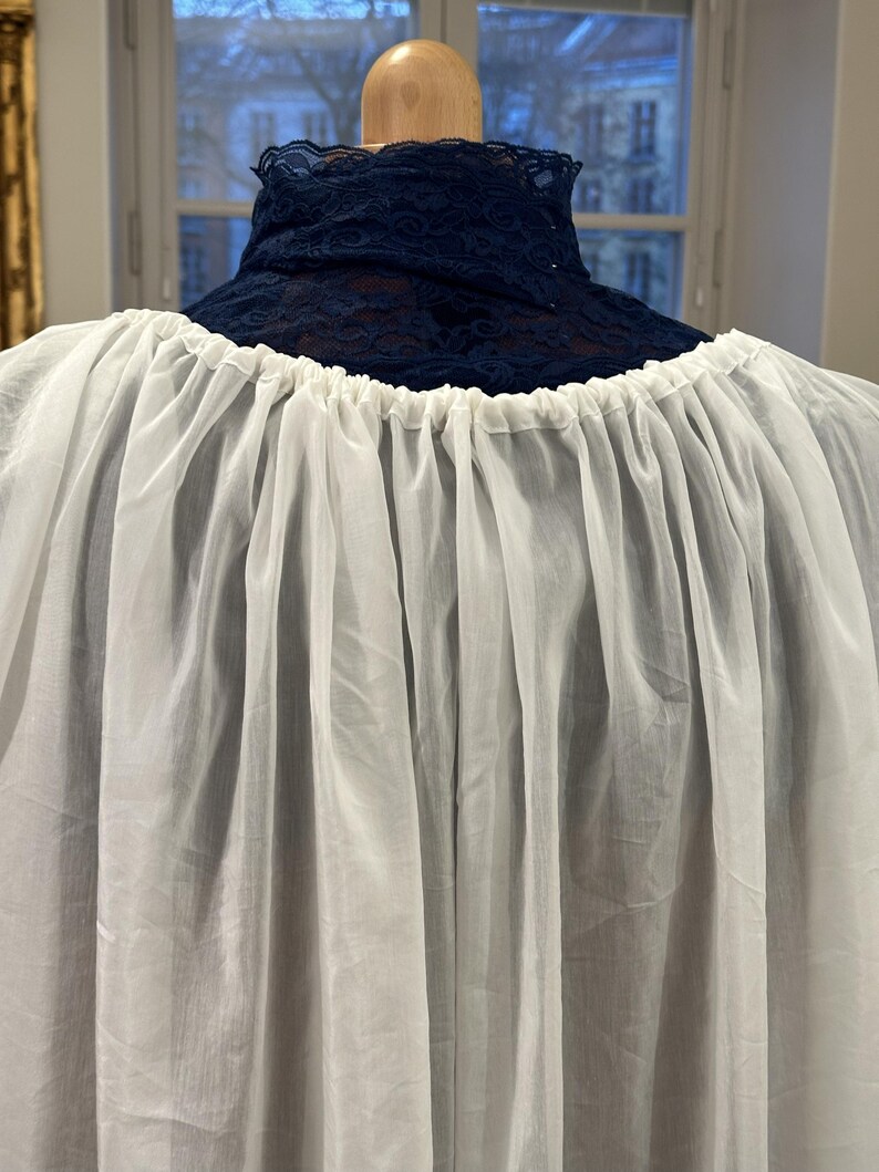 May include: A white, gathered fabric garment with a high, navy blue lace collar. The garment is displayed on a wooden mannequin head, showcasing the texture and drape of the fabric. The background includes a window with a view of buildings.