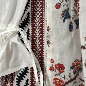 May include: Close-up of a white garment with patterned panels. The panels feature a mix of designs, including a black zigzag pattern, a brown and white leaf-like border, and a floral print with red and blue flowers. White ties are visible.