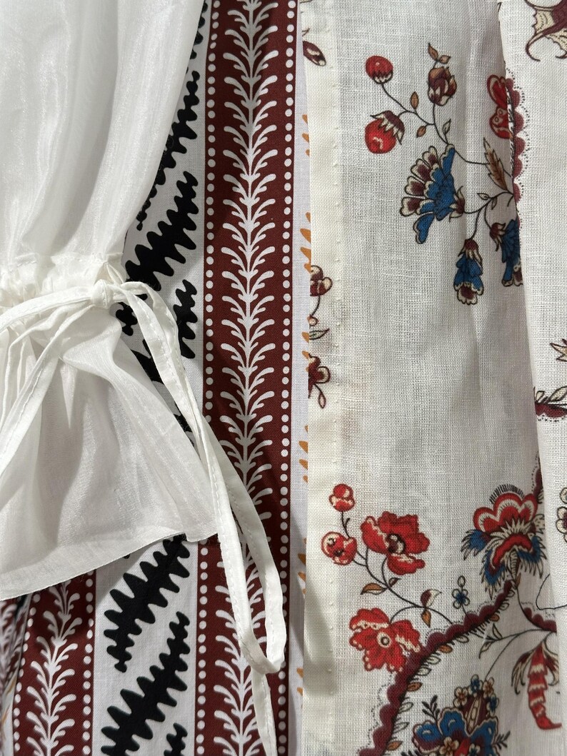 May include: Close-up of a white garment with patterned panels. The panels feature a mix of designs, including a black zigzag pattern, a brown and white leaf-like border, and a floral print with red and blue flowers. White ties are visible.