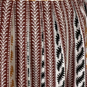 May include: Close-up of a pleated skirt with a repeating pattern. The skirt features vertical stripes in brown, white, and black. The brown stripes have a white dotted line and a white leaf design. The top edge has a zigzag pattern in gold.