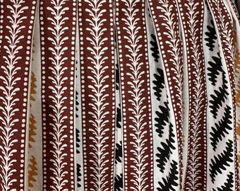 May include: Close-up of a pleated skirt with a repeating pattern. The skirt features vertical stripes in brown, white, and black. The brown stripes have a white dotted line and a white leaf design. The top edge has a zigzag pattern in gold.