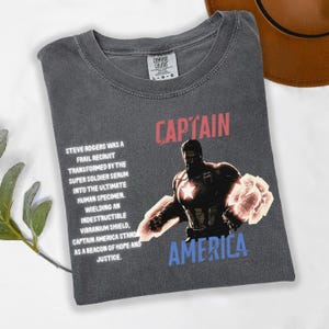 May include: Dark gray t-shirt featuring Captain America with the text "CAPTAIN AMERICA". The shirt includes a graphic of Captain America and text describing his origin and powers. Made by Comfort Colors.