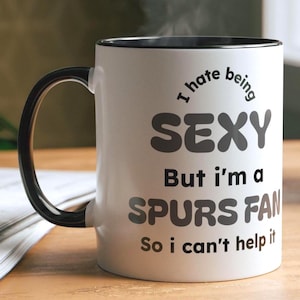 May include: White ceramic mug with a black handle and rim. The mug features the text "I hate being SEXY But I'm a SPURS FAN So i can't help it" in black and gray lettering. The mug is on a wooden surface.