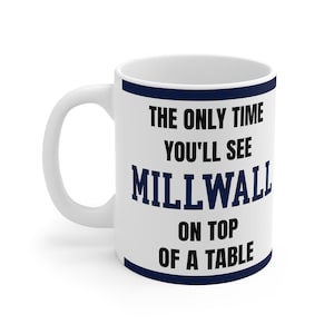 Pode incluir: Caneca de cerâmica branca com borda e alça azul marinho. A caneca apresenta o texto "THE ONLY TIME YOU'LL SEE MILLWALL ON TOP OF A TABLE" em letras pretas e azul marinho.