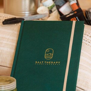 Könnte beinhalten: Ein dunkelgrünes Notizbuch mit den Worten "SALT THERAPY SURF JOURNAL" in goldfarbenen Buchstaben. Das Notizbuch hat ein cremefarbenes Gummiband und ein kleines Logo mit Sonne und Wellen. Pinsel und Kunstbedarf sind im Hintergrund.