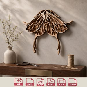 May include: A layered wooden moth wall art piece in shades of brown, mounted on a neutral-colored wall. The artwork features intricate cut-out details, creating a three-dimensional effect. The moth's wings are spread, and its body has a detailed design.