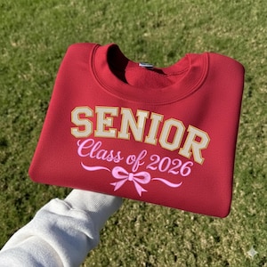 Op de afbeelding: Een rode sweatshirt met het woord "SENIOR" in gouden blokletters, "Class of 2026" in roze script en een roze strik. De sweatshirt wordt omhoog gehouden tegen een achtergrond van groen gras.