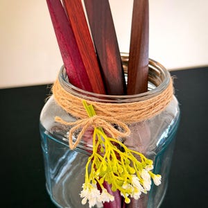 May include: A clear glass jar decorated with twine and a yellow and white floral arrangement. Inside the jar are several wooden tools in shades of red and brown. The jar is on a black surface.