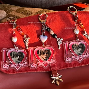 Puede incluir: Tres llaveros con colgantes en forma de corazón que dicen "I Love My Boyfriend". Cada llavero tiene una foto de una persona dentro del corazón, un pequeño dije de pistola y cuentas decorativas. Los llaveros cuelgan de un bolso rojo.