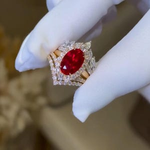 May include: A gold ring with a large oval ruby center stone, surrounded by smaller diamonds. The ring has a halo design and intricate detailing. The ruby is a deep red color, and the diamonds sparkle. The ring is held by a gloved hand.