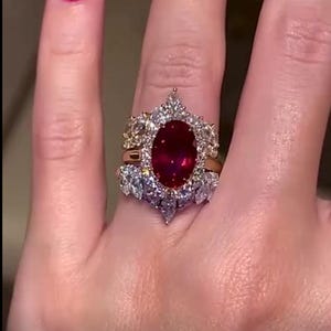 May include: A gold ring with a large oval ruby center stone, surrounded by smaller diamonds. The ring has a detailed, ornate design with additional diamond accents. The ruby is a deep red color, and the diamonds sparkle.