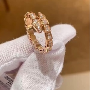 Puede incluir: Un anillo de serpiente de oro rosa con detalles de diamantes. El anillo está diseñado para parecerse a una serpiente, con la cabeza y la cola uniéndose para formar un círculo completo. El anillo se muestra sobre una superficie blanca.