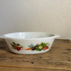 May include: White oval serving bowl with a colourful vegetable design. The bowl has two small handles and is decorated with illustrations of radishes, carrots, aubergine, and other vegetables. The bowl is on a wooden surface.