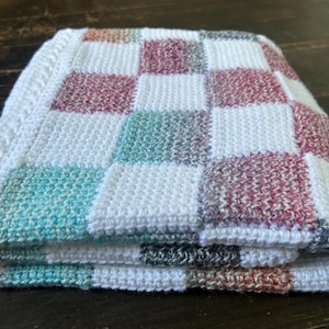 Op de afbeelding: Een gevouwen, handgebreide deken met een dambordpatroon. De vierkanten wisselen af tussen wit, blauwgroen, bordeauxrood en grijs. De deken is gemaakt van zacht garen en heeft een getextureerd uiterlijk. De deken is netjes opgevouwen.