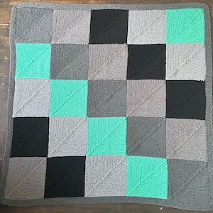 May include: A square, knitted blanket with a geometric pattern. The design features squares in shades of gray, black, and teal. The blanket has a gray border and is likely made of soft yarn, suitable for a baby or as a decorative throw.