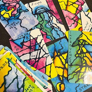 Può includere: Una collezione di carte dei tarocchi colorate con disegni astratti e testo scritto a mano. Le carte presentano tonalità vibranti di blu, rosa, giallo e verde, con parole come "Sacerdotessa", "Coppe" e "Miglioramento". L'opera d'arte ha uno stile unico e artistico.