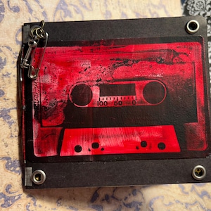 Op de afbeelding: Een zwart, rechthoekig cassettebandontwerp met een levendige rode overlay. De tape heeft een gedrukte schaal en is vastgezet met metalen klinknagels en een veiligheidsspeld. Het ontwerp roept een vintage esthetiek op.