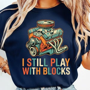 Peut inclure: T-shirt bleu marine avec une illustration de moteur de style vintage aux couleurs orange, turquoise et argent. Le texte "I STILL PLAY WITH BLOCKS" est imprimé en dessous en orange, turquoise et beige.