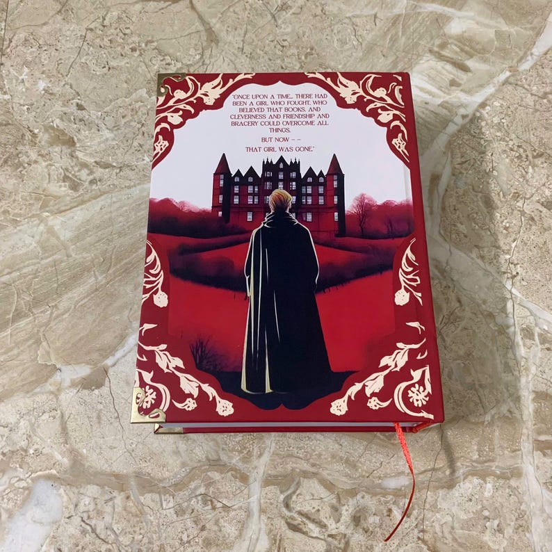 K&ouml;nnte beinhalten: Ein rotes Hardcover-Buch mit goldenen Kanten. Die Illustration auf dem Einband zeigt eine Person in einem schwarzen Umhang vor einem Schloss. Der Text auf dem Einband lautet "ONCE UPON A TIME...THAT GIRL WAS GONE."