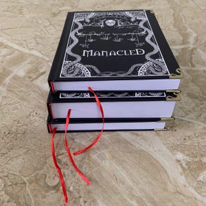 Pu&ograve; includere: Una pila di tre libri con copertina rigida nera con la scritta "MANACLED" in bianco. Le copertine presentano illustrazioni bianche ornate e angoli dorati. Segnalibri a nastro rosso sporgono dalle pagine.