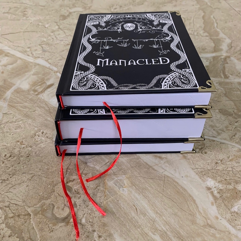 Pu&ograve; includere: Una pila di tre libri con copertina rigida nera con la scritta "MANACLED" in bianco. Le copertine presentano illustrazioni bianche ornate e angoli dorati. Segnalibri a nastro rosso sporgono dalle pagine.