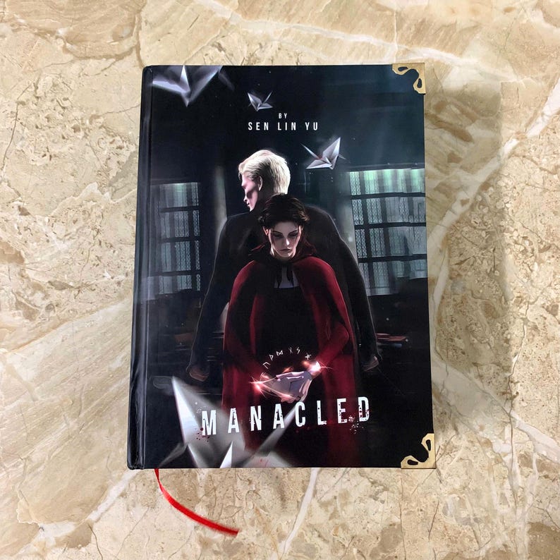 K&ouml;nnte beinhalten: Ein Hardcover-Buch mit dem Titel "MANACLED" von Sen Lin Yu. Das Cover zeigt eine dunkle Illustration von zwei Figuren, eine in einem roten Umhang, mit geometrischen Formen und einem roten Lesezeichen. Goldene Eckensch&uuml;tzer sind sichtbar.