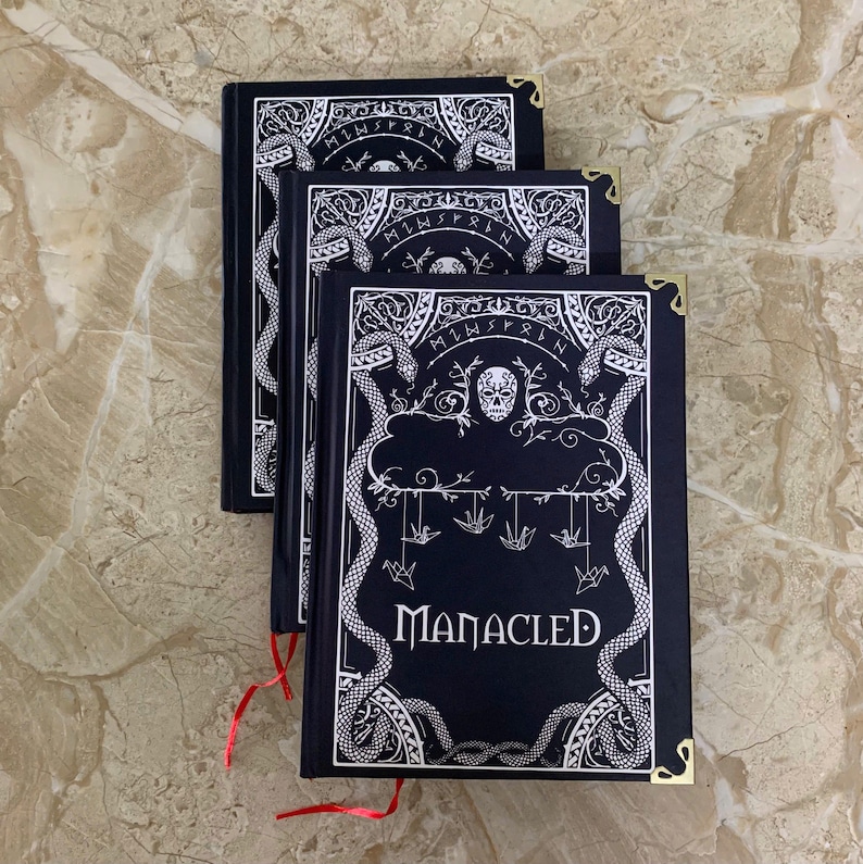 Pu&ograve; includere: Tre libri con copertina rigida neri con intricati disegni bianchi, tra cui serpenti, un teschio e la parola "MANACLED". Ogni libro ha protezioni angolari dorate e un segnalibro a nastro rosso. I libri sono impilati su una superficie chiara.