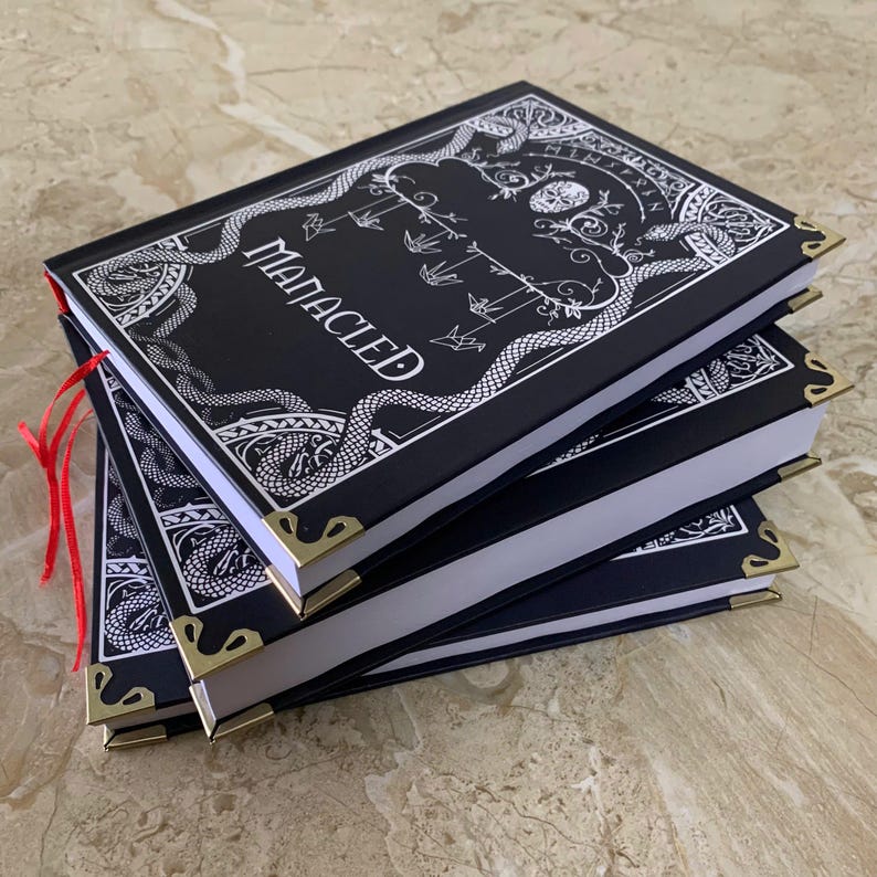 Pu&ograve; includere: Una pila di diari neri con protezioni angolari dorate. La copertina del diario superiore presenta la parola "MANACLED" in scrittura bianca, insieme a un disegno a linee bianche di serpenti, uccelli e altri simboli. Un segnalibro a nastro rosso &egrave; visibile.