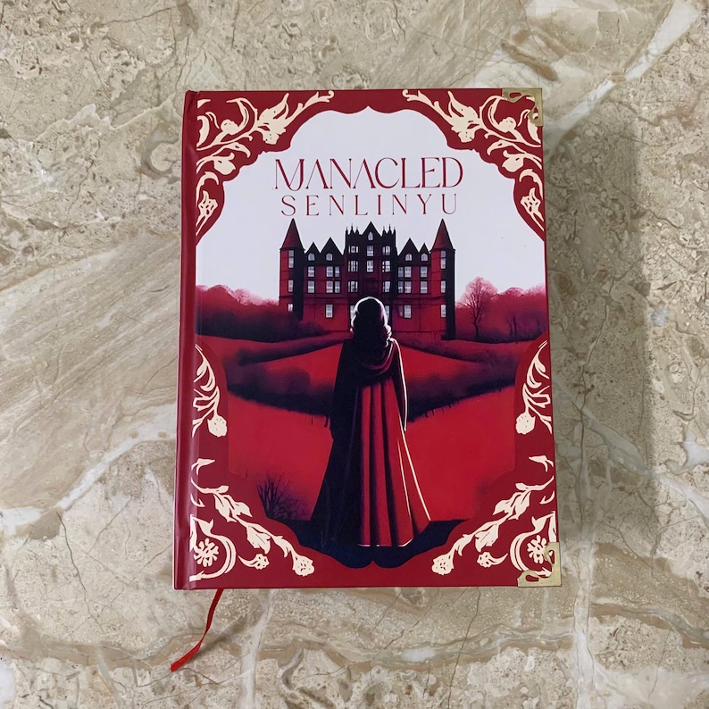 K&ouml;nnte beinhalten: Ein Hardcover-Buch mit dem Titel "MANACLED SENLINYU" mit rotem Einband. Das Cover zeigt eine Schlossillustration und eine Figur in einem roten Umhang. Dekorative florale Elemente und ein rotes Lesezeichen sind ebenfalls zu sehen.
